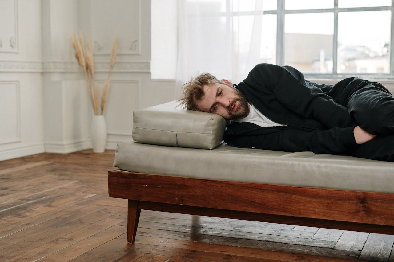 A bearded man lying on a couch in a bright, minimalistic room, looking contemplative.